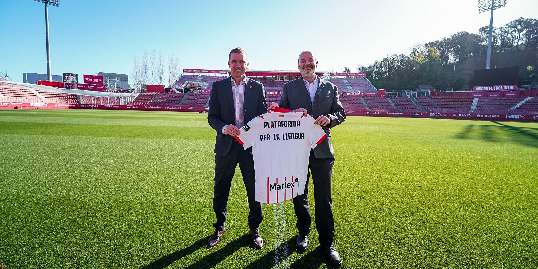 El Girona FC ha signat un conveni de col·laboració amb Plataforma per la Llengua per fomentar i normalitzar l’ús del català dins del club. L’acord, rubricat pels presidents Delfí Geli i Òscar Escuder a l’estadi de Montilivi, contempla diverses línies d’actuació per reforçar la presència de la llengua en tots els àmbits de l’entitat.
Entre les mesures previstes hi ha l’avaluació de la situació sociolingüística als equips de futbol base, el foment de l’aprenentatge del català entre tècnics i jugadors estrangers, així com la promoció de vocabulari específic del sector esportiu. A més, el pla també posa el focus en reforçar la presència del català en la comunicació del club, especialment a les xarxes socials, i en incentivar que els jugadors del primer equip utilitzin la llengua en les seves intervencions públiques. També es treballarà amb els patrocinadors perquè integrin el català en la seva activitat.
Per tal de garantir l’eficàcia del projecte, es durà a terme una diagnosi de la situació lingüística al futbol base, que permetrà dissenyar accions concretes basades en dades objectives. En aquest sentit, Plataforma per la Llengua oferirà assessorament al club per elaborar un protocol d’ús del català i desenvolupar activitats formatives en matèria de sensibilització lingüística. Aquestes iniciatives comptaran amb la col·laboració d’institucions locals com l’Ajuntament de Girona i el Consorci per a la Normalització Lingüística.
El president del Girona, Delfí Geli, ha destacat que aquest acord reafirma el compromís del club amb la llengua i el territori, amb la voluntat de convertir-se en un referent social i cultural. Per la seva banda, Òscar Escuder ha subratllat la importància que un club de Primera Divisió impulsi l’ús del català, especialment en àmbits com l’esport i el lleure, i ha expressat el desig que aquesta iniciativa serveixi d’exemple per a altres entitats esportives.